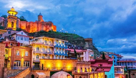 Georgian Delights Tbilisi Old Town at dusk with illuminated buildings nestled below Narikala Fortress on the hillside, Georgia